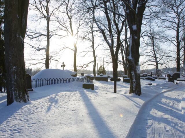 Luminen hautausmaa kylpee kirkkaassa auringonpaisteessa. Vanhat puut ja haudat lepäävät lumipeitteen alla.