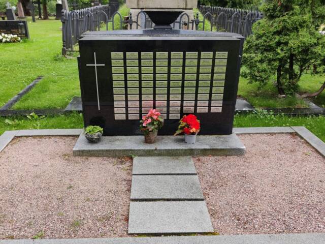 Muistokivi, jossa on pieniä metallisia laattoja. Laatoissa on ihmisten nimiä. Kiven edessä on muutama kukka.