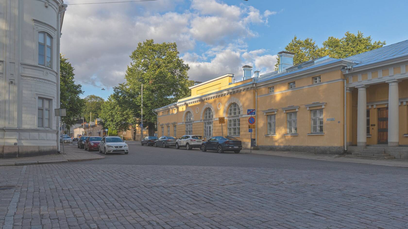 Keltainen talo Maaherran Makasiini. Edessä asfalttia ja pysäköityjä autoja. Taustalla vehreitä puita ja sinistä taivasta.