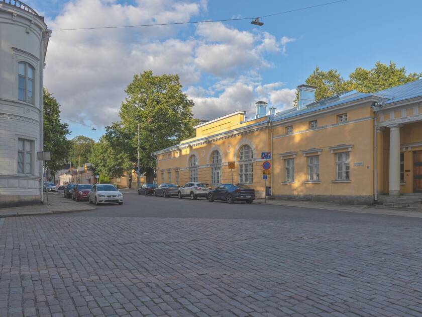 Keltainen talo Maaherran Makasiini. Edessä asfalttia ja pysäköityjä autoja. Taustalla vehreitä puita ja sinistä taivasta.
