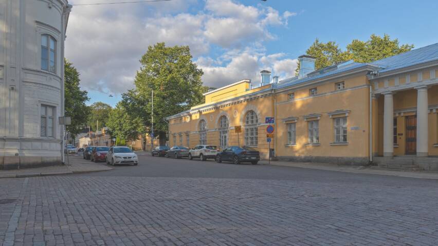 Keltainen talo Maaherran Makasiini. Edessä asfalttia ja pysäköityjä autoja. Taustalla vehreitä puita ja sinistä taivasta.