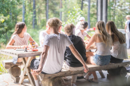 Nuoria istuu pöydän ympärillä terassilla. Nuoret katselevat taustalla olevaa pingispeliä.