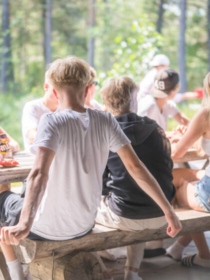 Nuoria istuu pöydän ympärillä terassilla. Nuoret katselevat taustalla olevaa pingispeliä.