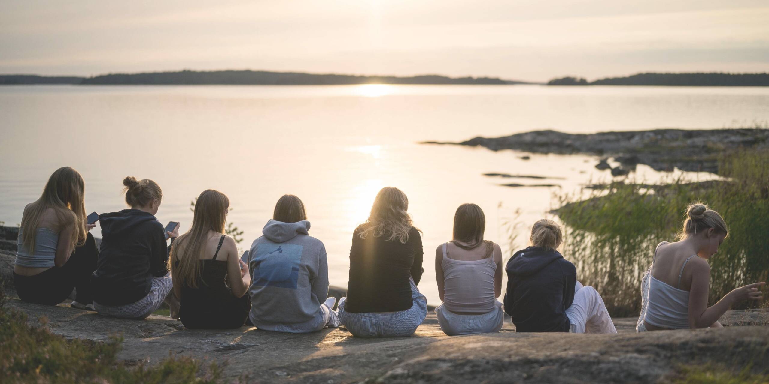 Rannalla istuvia nuoria naisia, jotka katsovat auringonlaskua järven yli. Taustalla rauhallinen maisema ja heijastuksia vedessä.