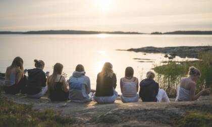 Rannalla istuvia nuoria naisia, jotka katsovat auringonlaskua järven yli. Taustalla rauhallinen maisema ja heijastuksia vedessä.