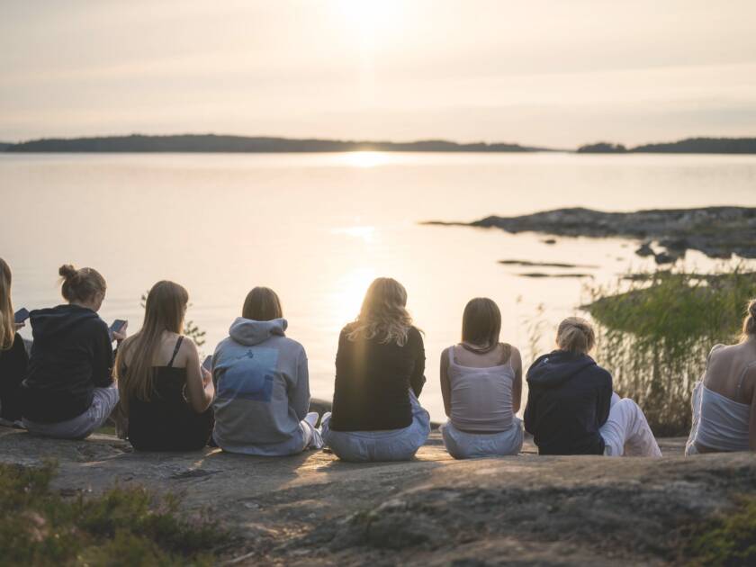 Rannalla istuvia nuoria naisia, jotka katsovat auringonlaskua järven yli. Taustalla rauhallinen maisema ja heijastuksia vedessä.