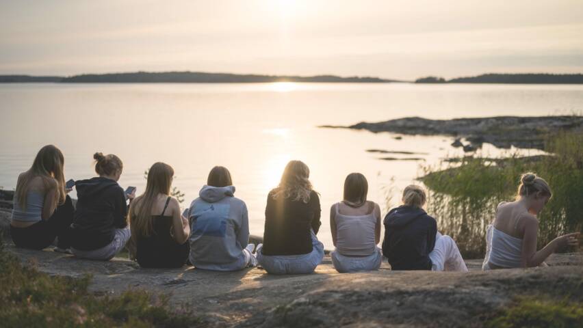 Rannalla istuvia nuoria naisia, jotka katsovat auringonlaskua järven yli. Taustalla rauhallinen maisema ja heijastuksia vedessä.