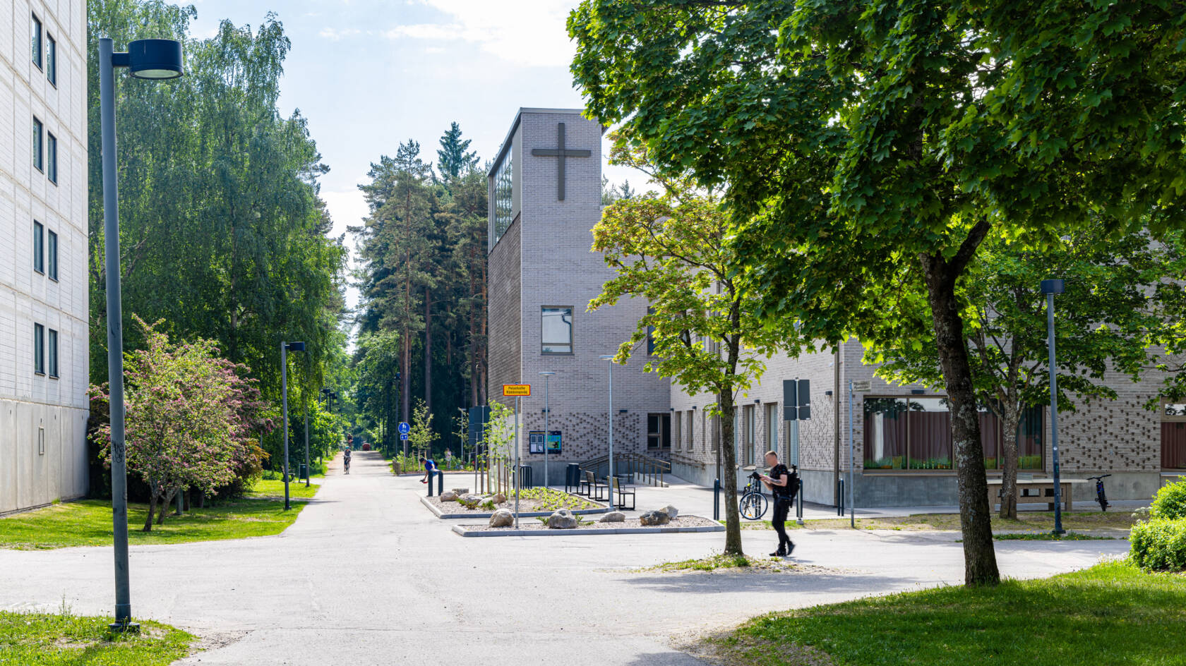 Stadsmiljö med en parkliknande gångväg, byggnader och gröna träd. I bakgrunden syns ett kors på byggnadens vägg. Heikki Raisanen RAEVEN VISUAL