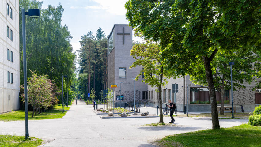 Kaupunkimaisema, jossa on puistomainen kävelytie, rakennuksia ja vihreitä puita. Taustalla näkyy risti rakennuksen seinässä. Heikki Raisanen RAEVEN VISUAL.