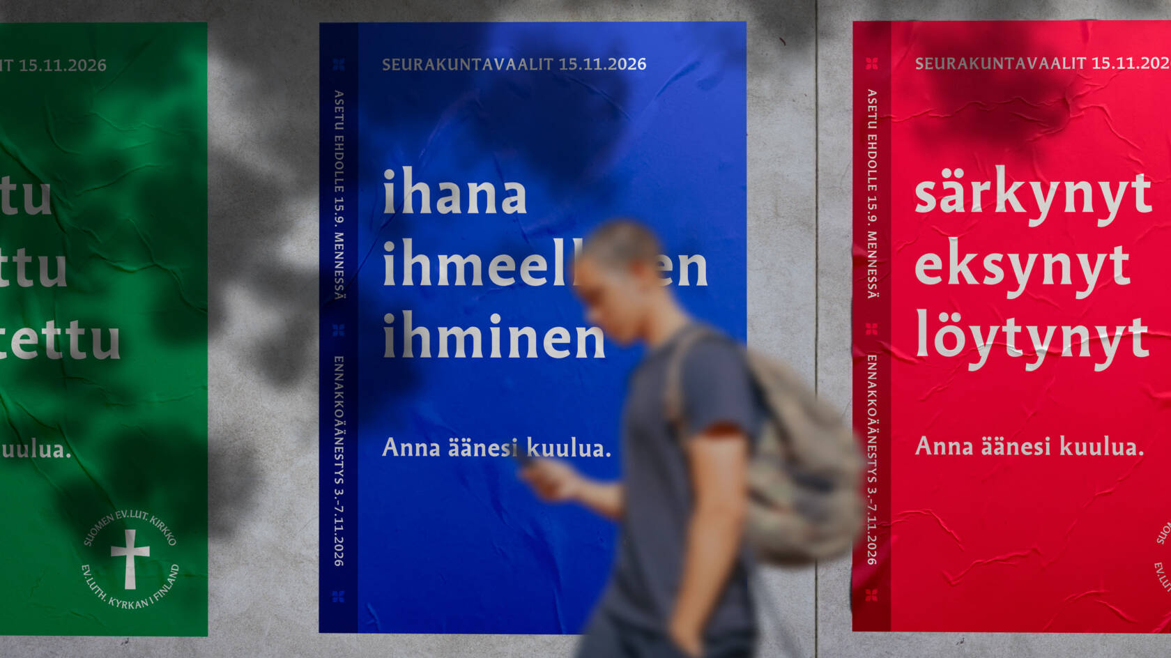 Kolme julistetta seinällä, joissa on eri värejä ja tekstejä. Henkilö kävelee ohi, taustalla näkyy punainen, sininen ja vihreä juliste.