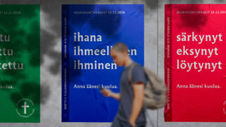 Kolme julistetta seinällä, joissa on eri värejä ja tekstejä. Henkilö kävelee ohi, taustalla näkyy punainen, sininen ja vihreä juliste.