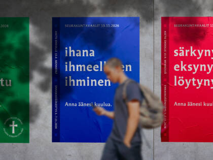 Kolme julistetta seinällä, joissa on eri värejä ja tekstejä. Henkilö kävelee ohi, taustalla näkyy punainen, sininen ja vihreä juliste.