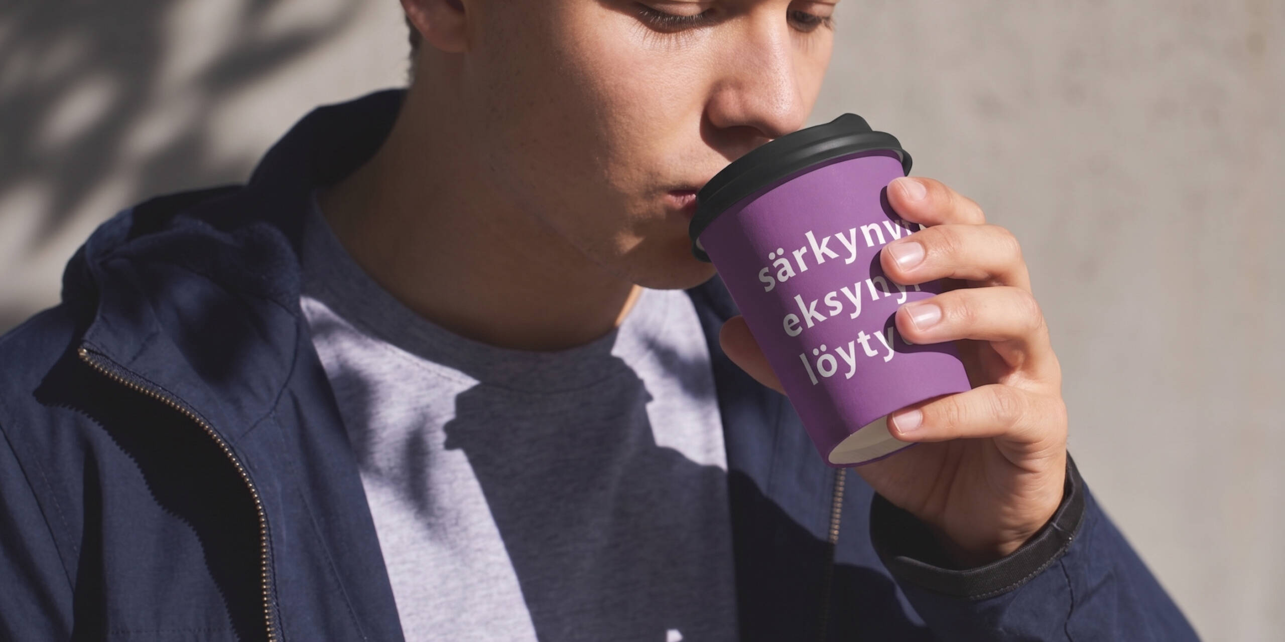 Mies juo kahvia violetista kupista, jossa on tekstiä. Hänellä on sininen takki ja harmaa paita.