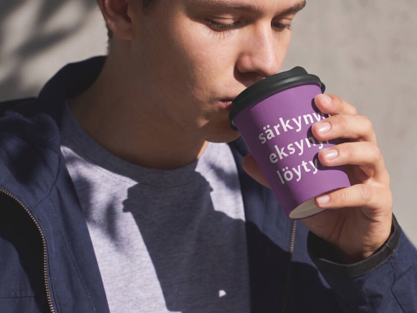 Mies juo kahvia violetista kupista, jossa on tekstiä. Hänellä on sininen takki ja harmaa paita.