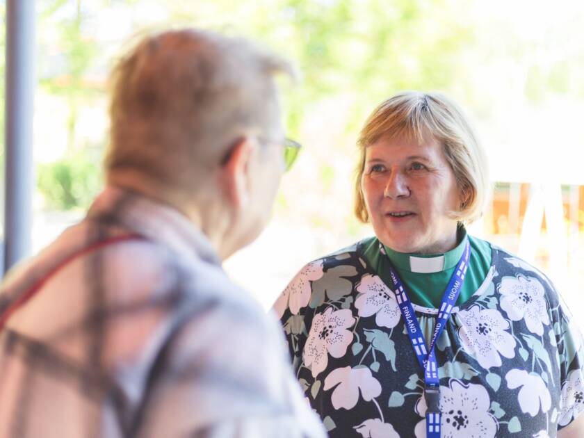 Kaksi henkilöä keskustelee ulkona, toinen pukeutunut diakonian tunnuksiin ja kukkakuosiseen puseroon. Taustalla näkyy vihreitä puita ja kirkas taivas.