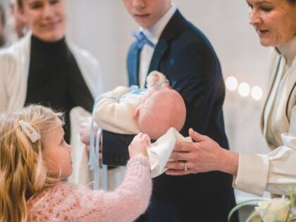 Pieni vauva on kastepuvussaan pojan sylissä. Pappi ja pieni tyttö pyyhkivät lapsen päätä pienellä kankaalla.