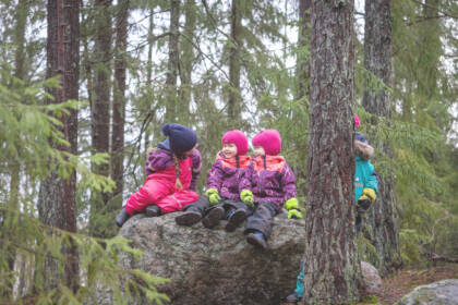 Kolme lasta istuu haalareissaan kivellä metsässä.