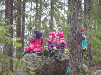 Kolme lasta istuu haalareissaan kivellä metsässä.
