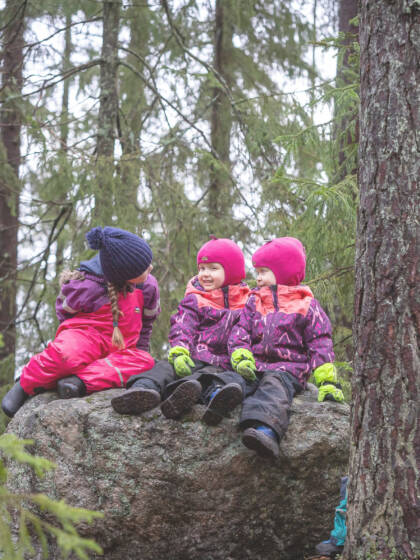 Kolme lasta istuu haalareissaan kivellä metsässä.