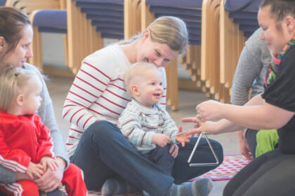 Vauva istuu äitinsä sylissä. Lastenohjaaja ojentaa triangelia vauvan eteen.