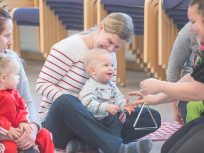 Vauva istuu äitinsä sylissä. Lastenohjaaja ojentaa triangelia vauvan eteen.