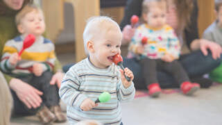 Vauva narskuttaa marakassia suussaan. Taustalla muita vauvoja äitiensä kanssa.