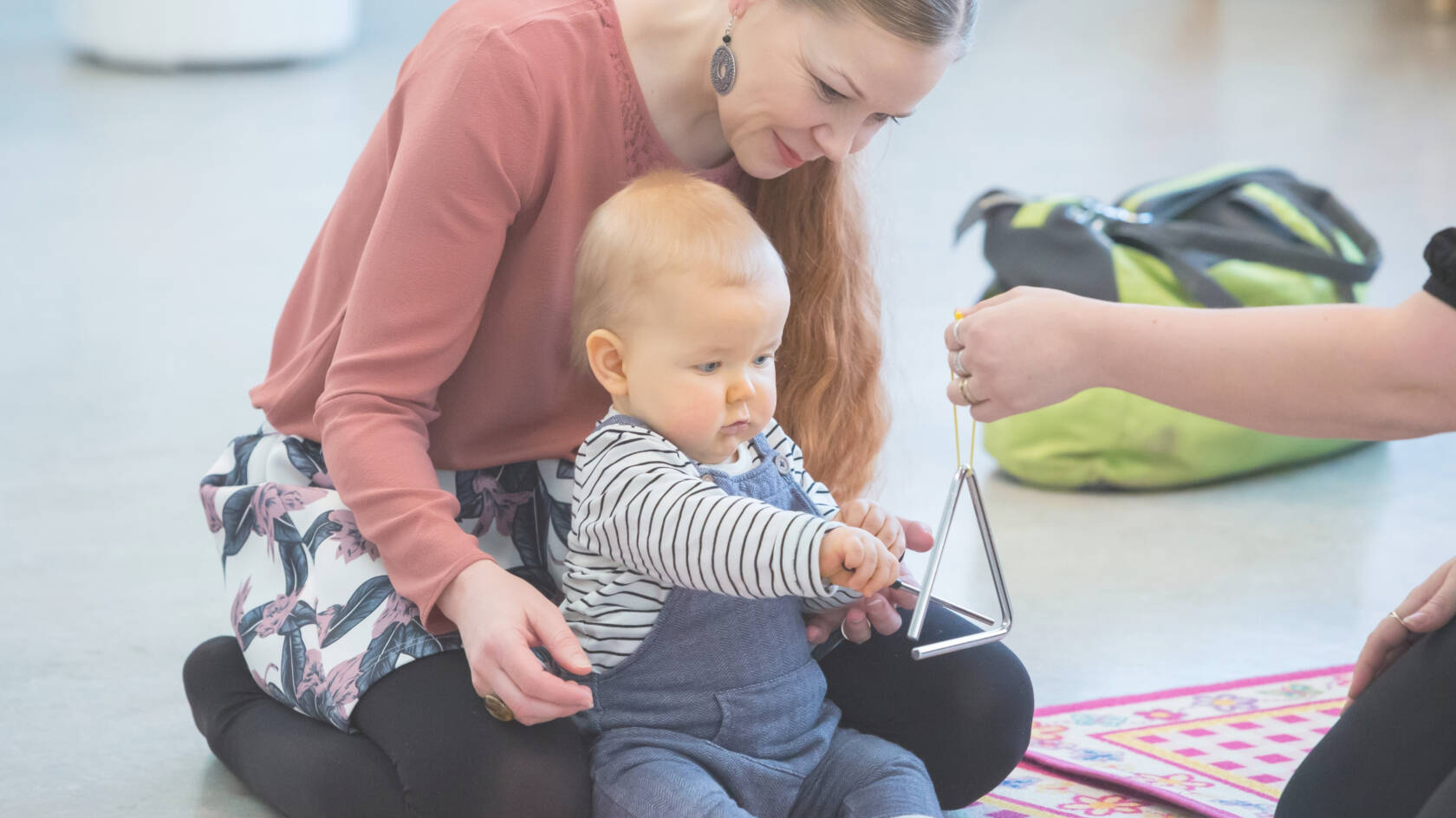 Vauva istuu äitinsä edessä lattialla. Käsi ojentaa triangelia vauvan eteen ja vauva kilauttaa sitä kapulalla.
