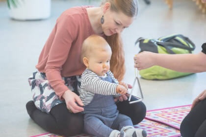 Vauva istuu äitinsä edessä lattialla. Käsi ojentaa triangelia vauvan eteen ja vauva kilauttaa sitä kapulalla.
