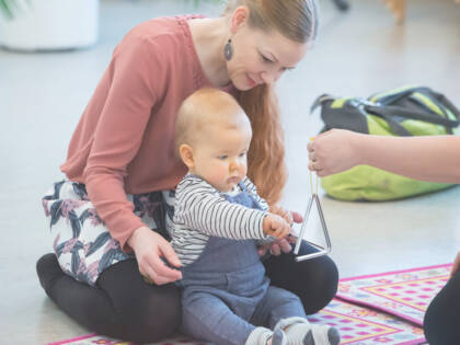 Vauva istuu äitinsä edessä lattialla. Käsi ojentaa triangelia vauvan eteen ja vauva kilauttaa sitä kapulalla.