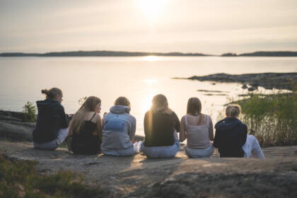 Nuoria istuu rivissä rantakalliolla. Aurinko on lasekmassa.