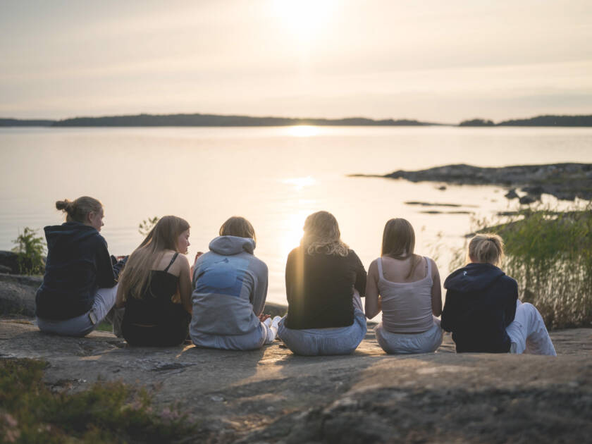 Nuoria istuu rivissä rantakalliolla. Aurinko on lasekmassa.