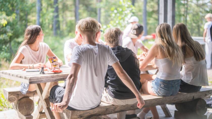 Nuoria istuu terassilla pöydän ääressä katselemassa muiden pingisten pelaamista. Pöydällä on karkkia ja muita tavaroita.
