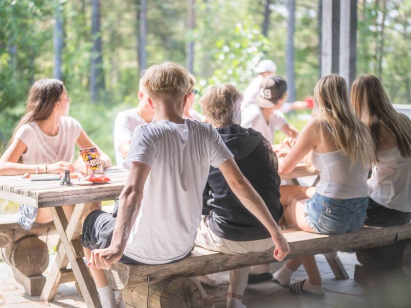 Nuoria istuu terassilla pöydän ääressä katselemassa muiden pingisten pelaamista. Pöydällä on karkkia ja muita tavaroita.