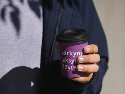 En man håller en lila kaffekopp med texten "särkynyt eksynyt löytö". I bakgrunden syns skuggor och en ljus yta.