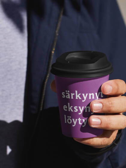 Mies pitää violetista kahvikupista, jossa on teksti "särkynyt eksynyt löytö". Taustalla näkyy varjoja ja vaaleaa pintaa.