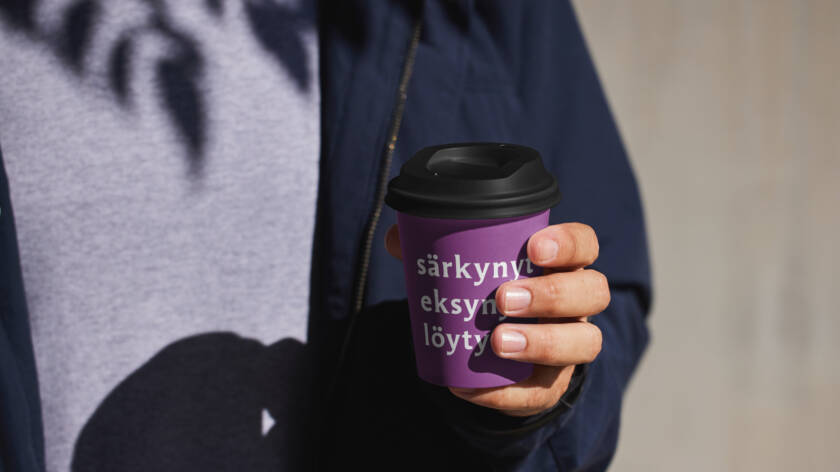 Mies pitää violetista kahvikupista, jossa on teksti "särkynyt eksynyt löytö". Taustalla näkyy varjoja ja vaaleaa pintaa.