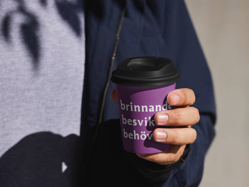 Mies pitää violetista kahvikupista, jossa on teksti "brinnande besvikna behov". Taustalla on vaaleanharmaa seinä ja varjoja.