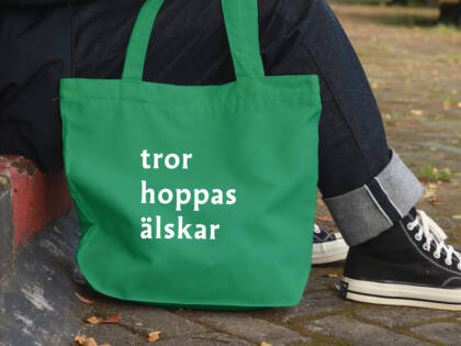 En grön tygväska med vita texter "tror", "hoppas" och "älskar". Väskan ligger bredvid en fot, med en stenlagd väg i bakgrunden.