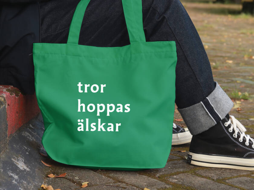 En grön tygväska med vita texter "tror", "hoppas" och "älskar". Väskan ligger bredvid en fot, med en stenlagd väg i bakgrunden.