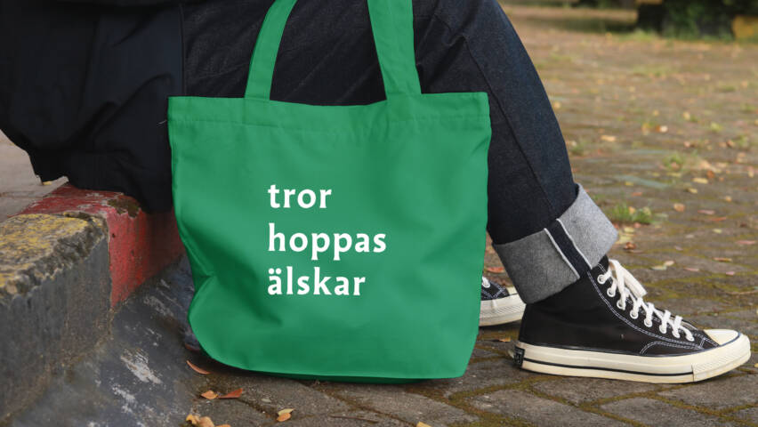 En grön tygväska med vita texter "tror", "hoppas" och "älskar". Väskan ligger bredvid en fot, med en stenlagd väg i bakgrunden.