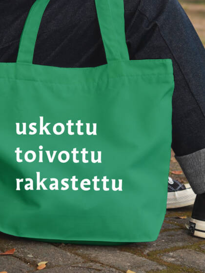 Vihreä kangaskassi, jossa on valkoiset sanat "uskottu", "toivottu" ja "rakastettu". Taustalla henkilö istuu kivetyksellä.