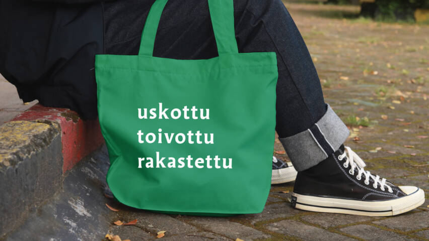 Vihreä kangaskassi, jossa on valkoiset sanat "uskottu", "toivottu" ja "rakastettu". Taustalla henkilö istuu kivetyksellä.