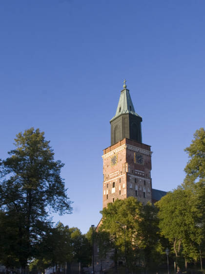Åbo domkyrkas torn mot en blå himmel, omgiven av gröna träd och den gamla kyrkobyggnaden.