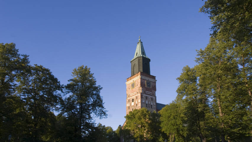 Turun tuomiokirkon torni sinistä taivasta vasten, ympärillä vihreitä puita ja kirkon vanha rakennus.