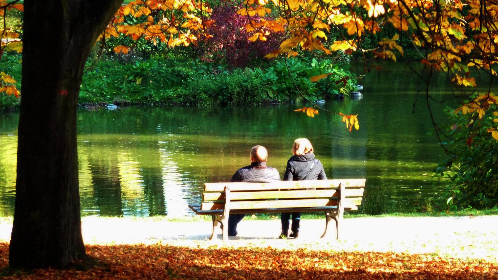 två vuxna som sitter på en parkbänk vid ån på hösten.