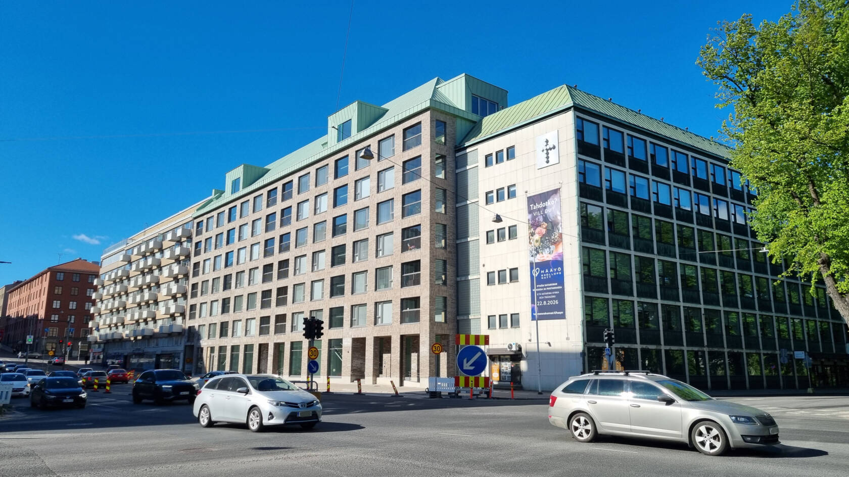 Kuvassa Aninkaistenkatu 1 uudisrakennus ja kulmittain sen vieressä seurakuntien toimitalo Eerikinkatu 3. Edessä kulkee autoja.