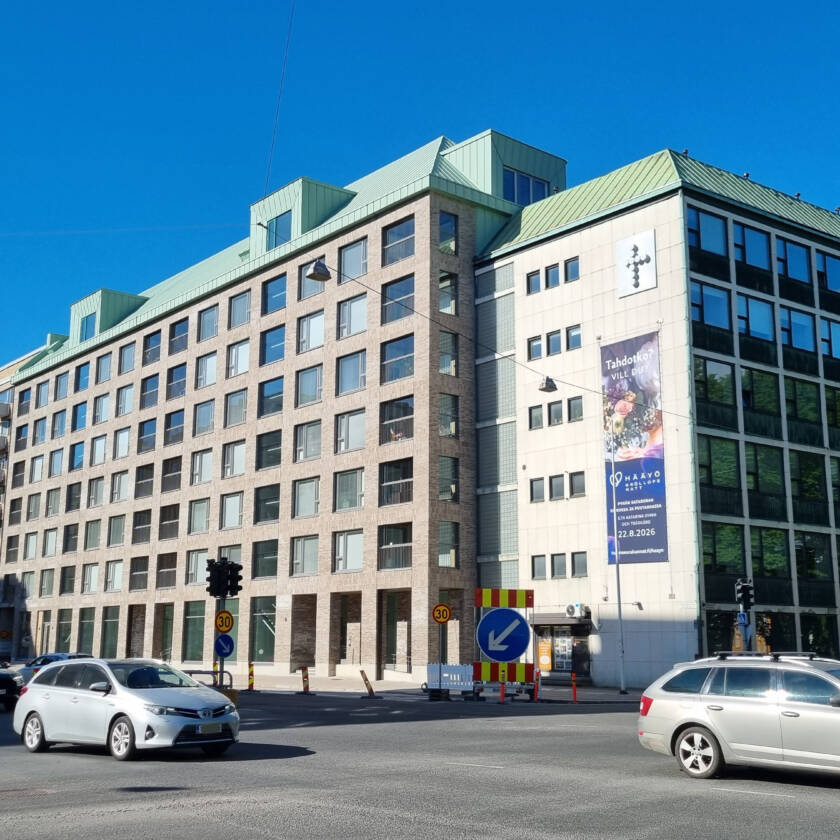 Kuvassa Aninkaistenkatu 1 uudisrakennus ja kulmittain sen vieressä seurakuntien toimitalo Eerikinkatu 3. Edessä kulkee autoja.