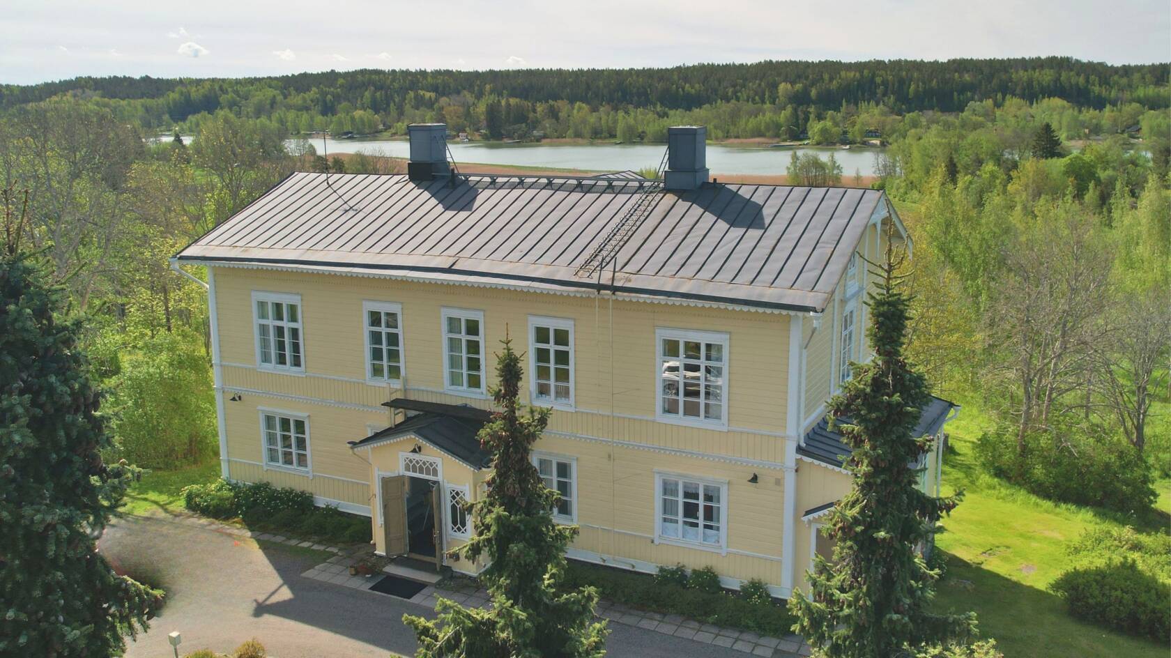 Keltainen suuri talo yläilmoista kuvattuna. Taustalla puita, peltoa ja meri.