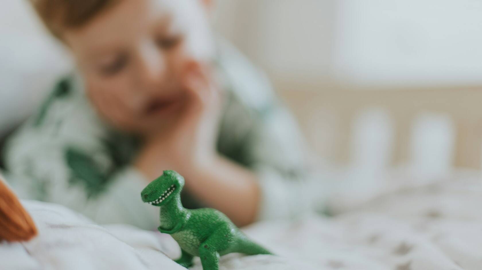 Sängyllä makoileva lapsi katsoo leludinosaurusta.