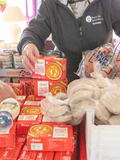 Pöydällä on suuri määrä sämpylöitä, valmiskeittopurkkeja, lihapiirakkapusseja ja makkaraa. Henkilö hupparissa ojentaa yhtä makkarapakettia ja linssikeittopurkkia.